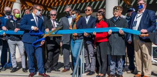 Multiple people standing outside of Mather Veterans Village, smiling and holding a blue ribbon, with another man standing in front with a large pair of blue scissors ready to cut the ribbon.