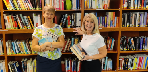 Two women organizing books on a book shelf, smiling towards the camera