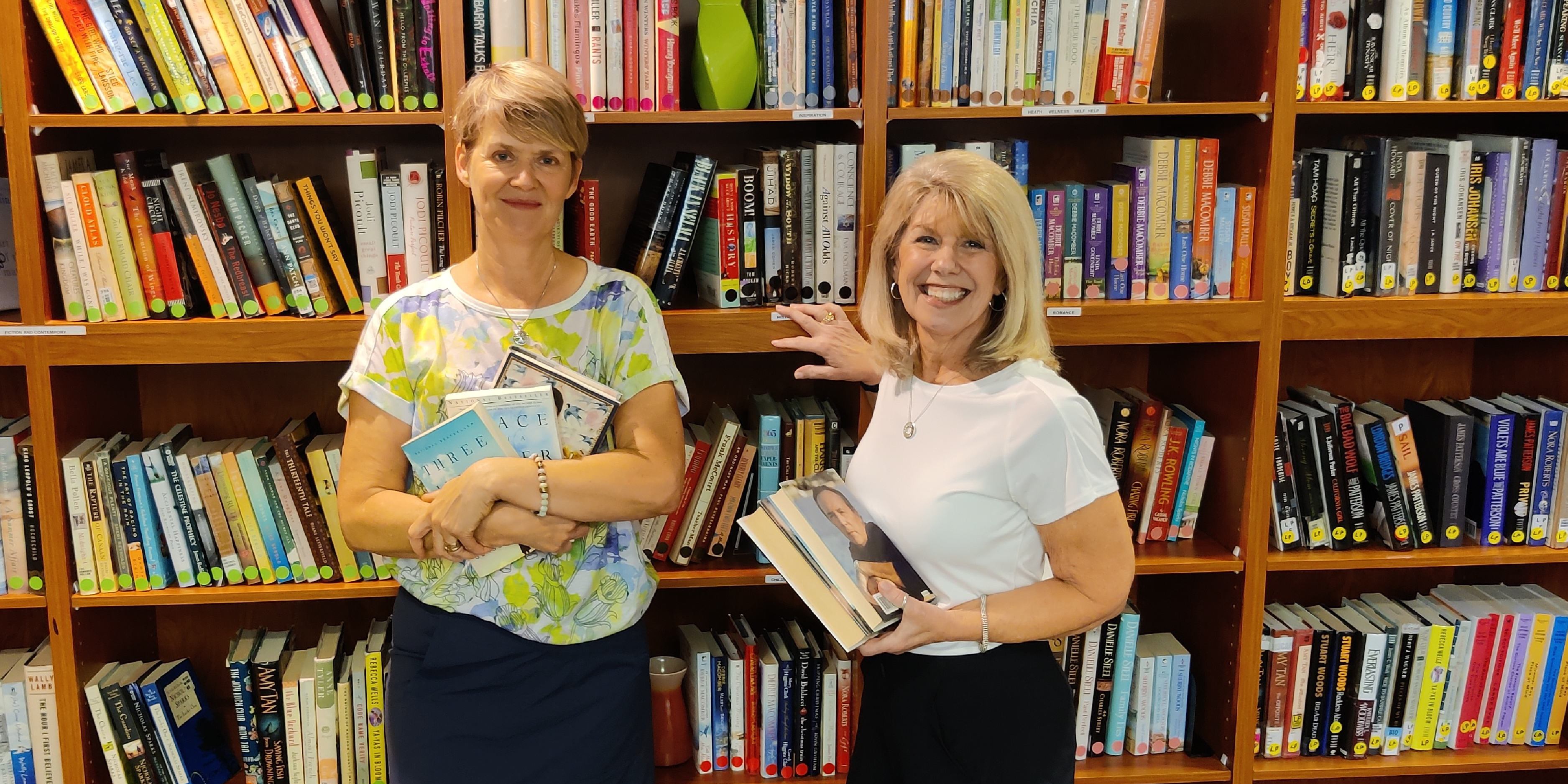 Two women organizing books on a book shelf, smiling towards the camera